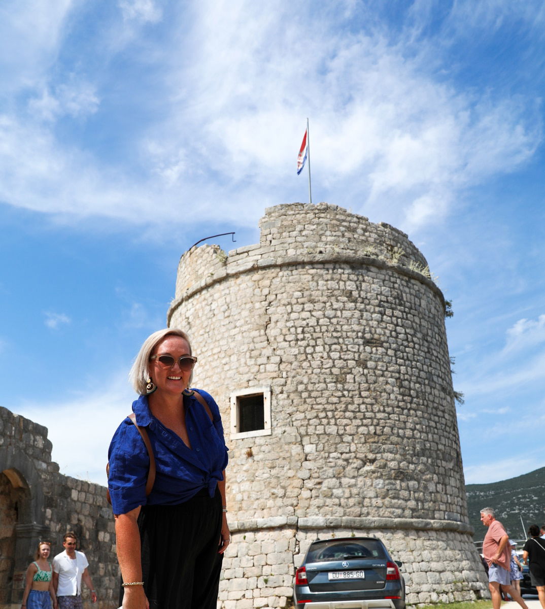 A smiling woman in a blue shirt stands in front of a stone tower with a flag on top, under the clear blue sky of Ston, Croatia. Several other people and a parked car can be seen in the background, capturing the charm of an Oyster Day trip.