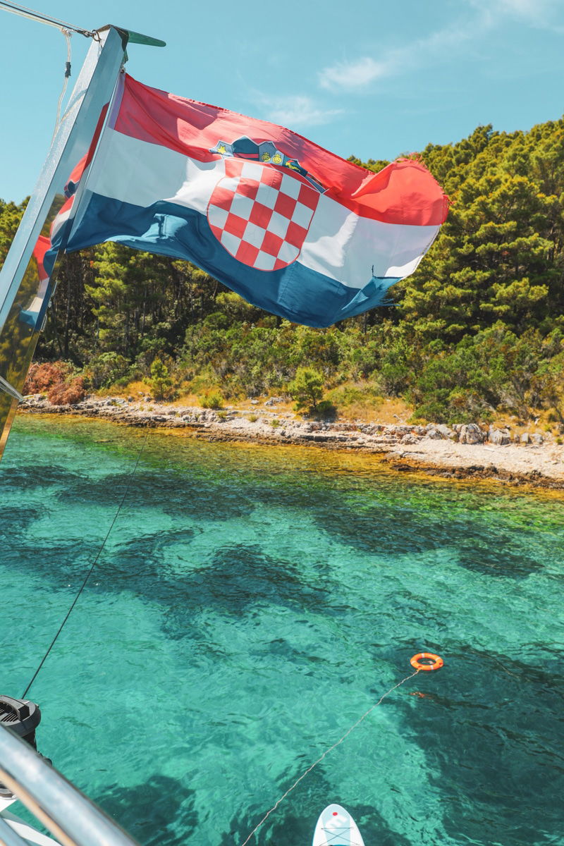 A Croatian flag waves on a boat with clear turquoise water and a wooded shoreline in the background, capturing the essence of swimming the Adriatic Sea.