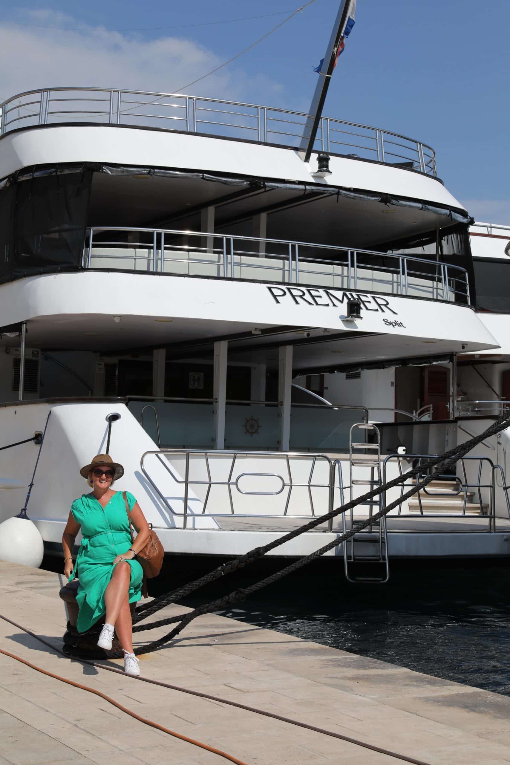 SJ in a green dress sits on a dock beside a large white yacht named "MS Premier" on a sunny day, ready to embark on their luxury cruise from Trogir.